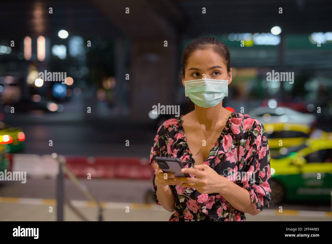 Donna che indossa maschera facciale per la protezione contro il coronavirus Covid-19 mentre in attesa di taxi di notte e utilizzando il telefono cellulare Foto Stock
