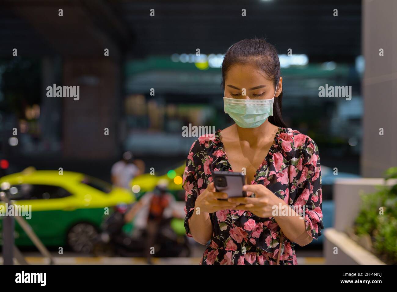 Donna che indossa maschera facciale per la protezione contro il coronavirus Covid-19 mentre in attesa di taxi di notte e utilizzando il telefono cellulare Foto Stock
