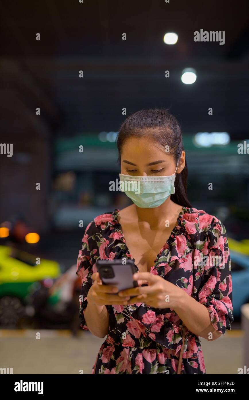 Donna che indossa maschera facciale per la protezione contro il coronavirus Covid-19 mentre in attesa di taxi di notte e utilizzando telefono cellulare verticale shot Foto Stock