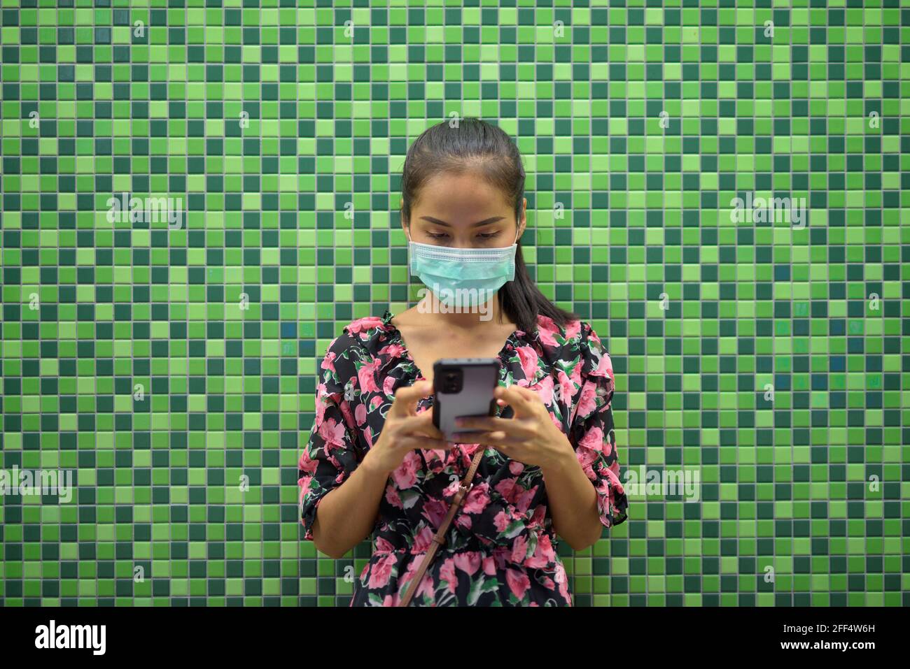 Ritratto di donna che indossa maschera per la protezione contro il coronavirus Covid-19 di notte contro parete colorata con spazio di copia mentre invio di sms con Foto Stock