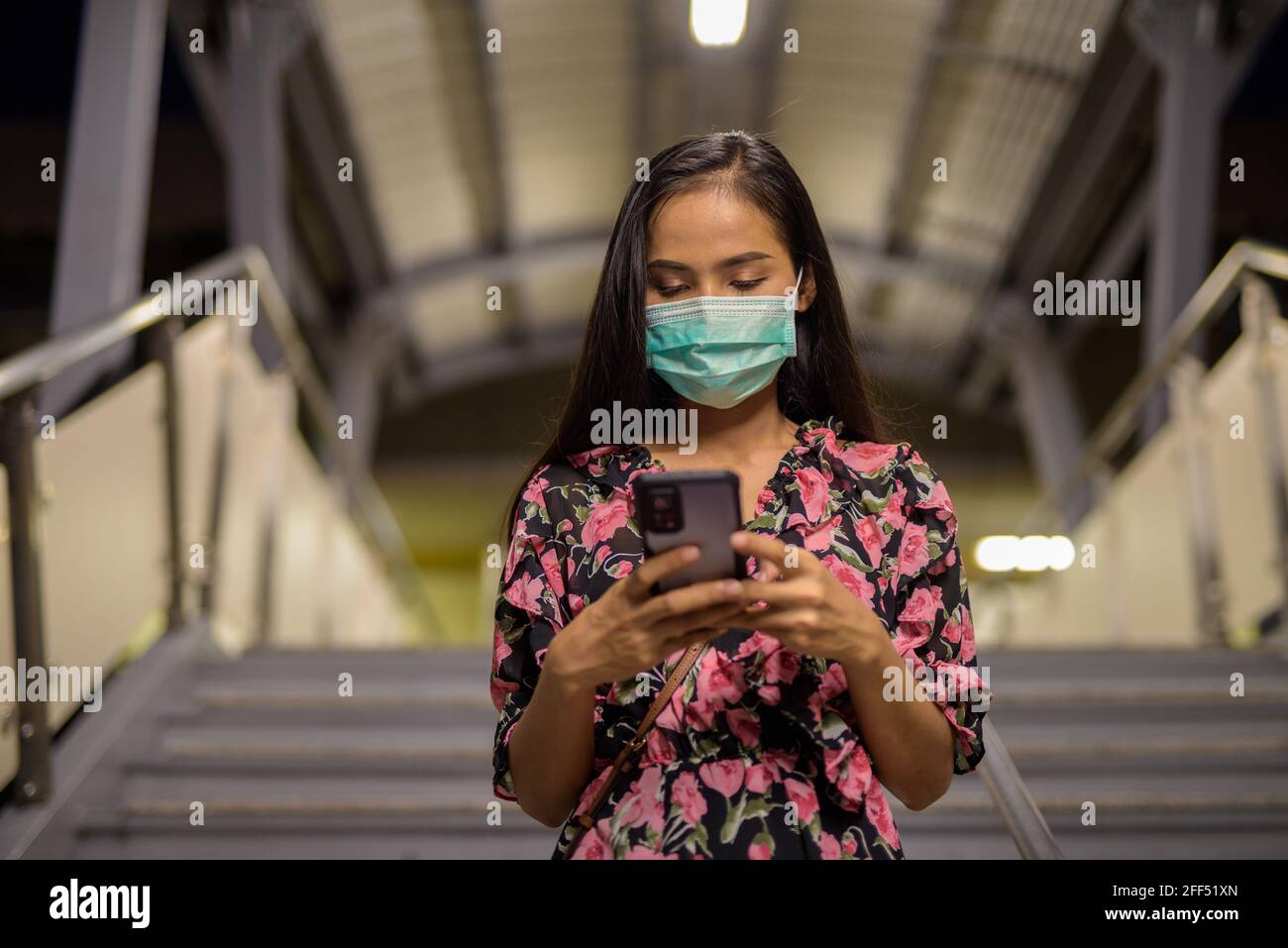 Ritratto di donna che indossa maschera per la protezione contro il coronavirus Covid-19 durante l'uso del telefono cellulare di notte Foto Stock