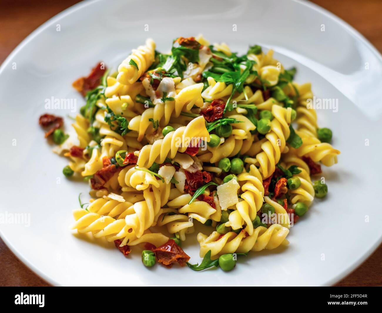 Fusilli con pomodori secchi, piselli, foglie di rucola e parmigiano su piatto bianco, primo piano. Foto Stock