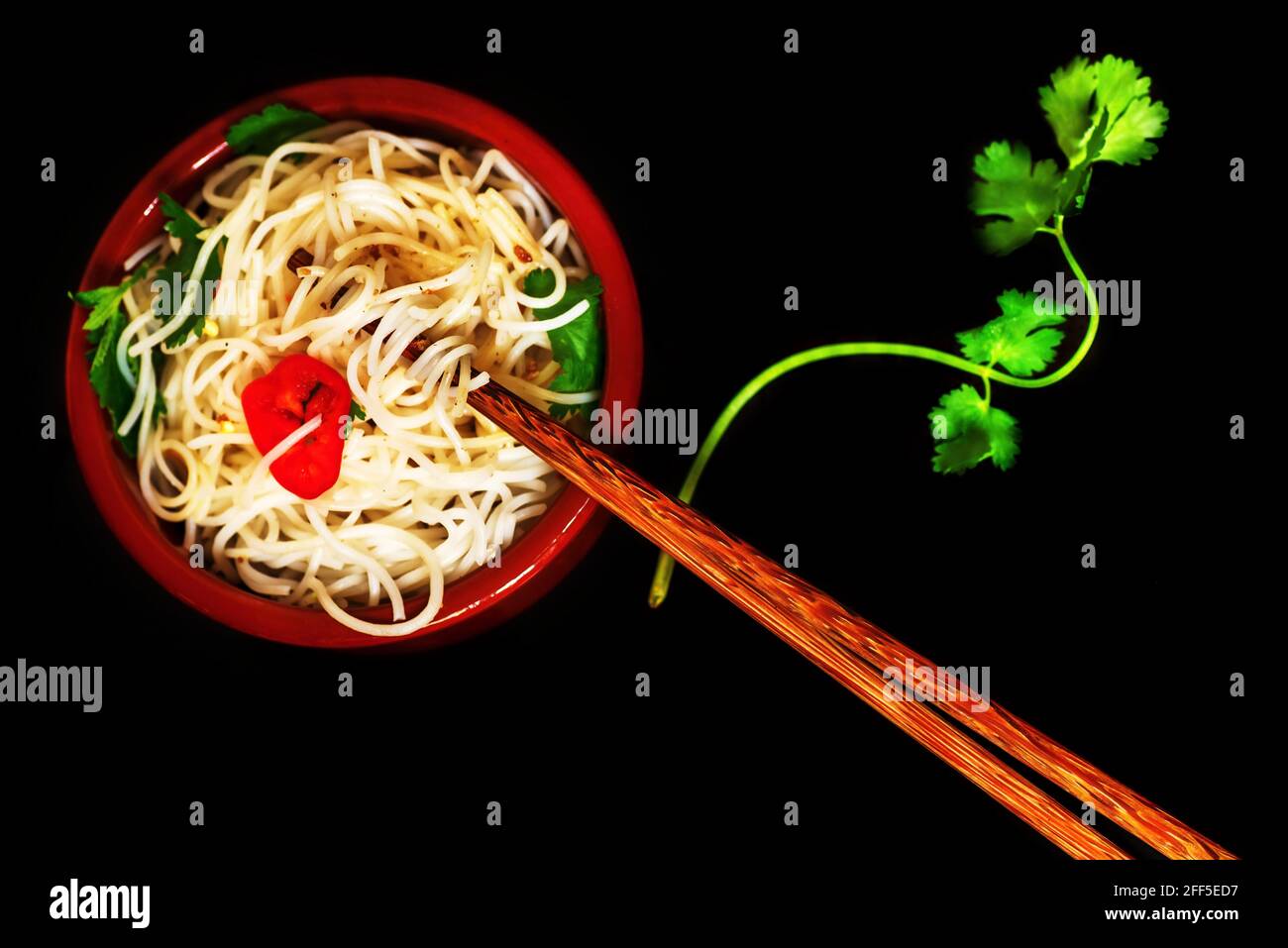 Ciotola in ceramica con tagliatelle bollite, erbe, peperoncino e pepe, foglie di coriandolo verde fresco e bastoncini a pezzetti su sfondo nero. Noodle asiatico. Foto Stock