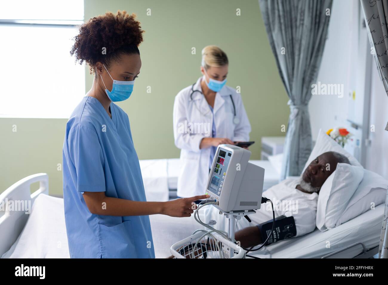 Razza mista dottore femminile che indossa maschera facciale utilizzando la pressione sanguigna monitor nella sala paziente Foto Stock