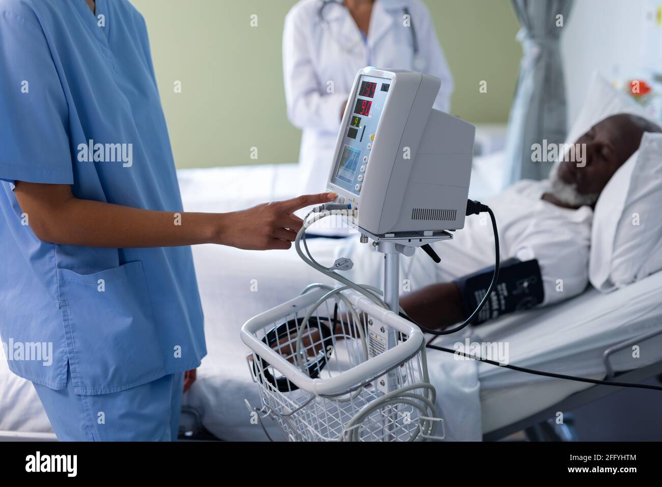 Mezza sezione del medico femminile di razza mista che usa il dispositivo di controllo della pressione sanguigna nella stanza del paziente Foto Stock
