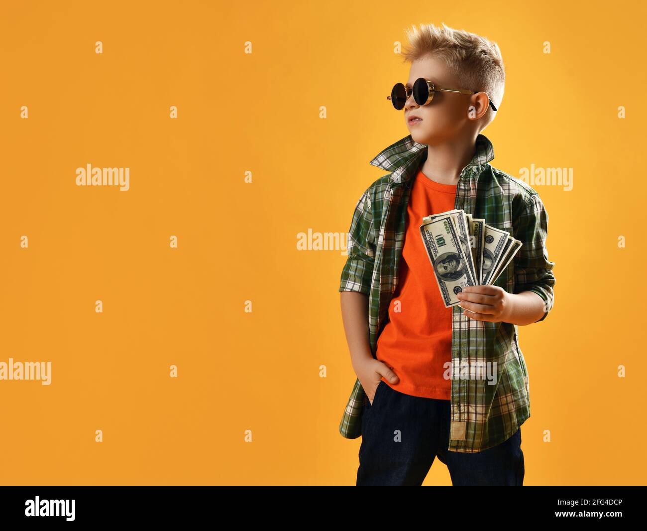 Infalliente ragazzo ricco audace in occhiali da sole rotondi, camicia a quadri e jeans stand reggendo bundle di contanti Foto Stock