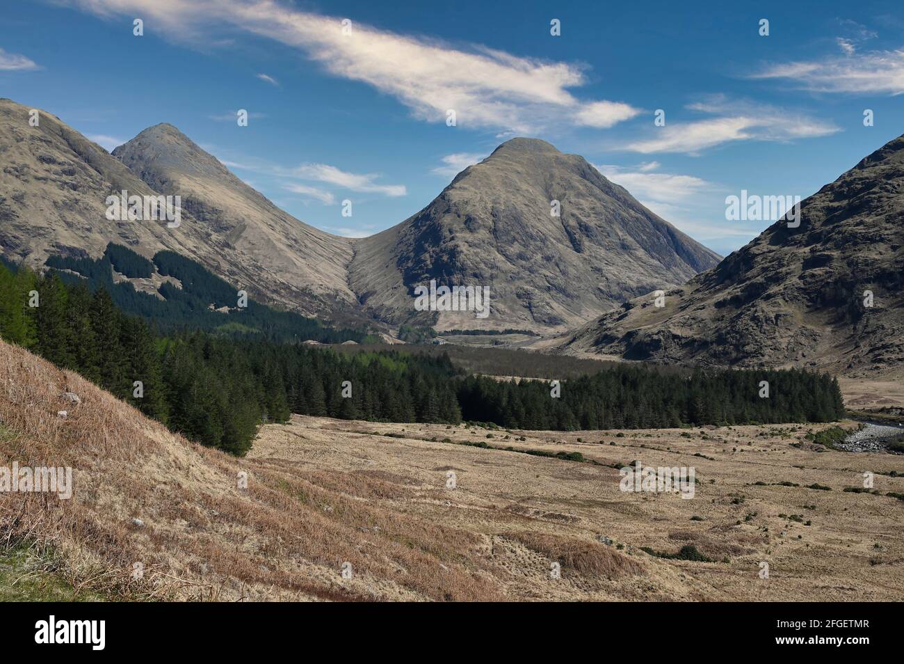 Montagne di Glen Etive nelle Highlands scozzesi in una giornata di sole con cielo blu e nuvole di luce. Valle e foresta di fronte alle montagne. Foto Stock