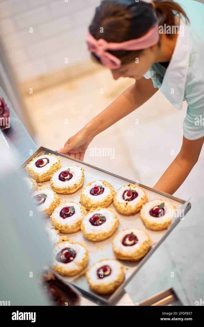 Una giovane piccola impresa donna proprietario sta preparando le sue deliziose ciambelle fatte a mano di aspetto irresistibile per una pasticceria. Pasticceria, dessert, dolce, m Foto Stock