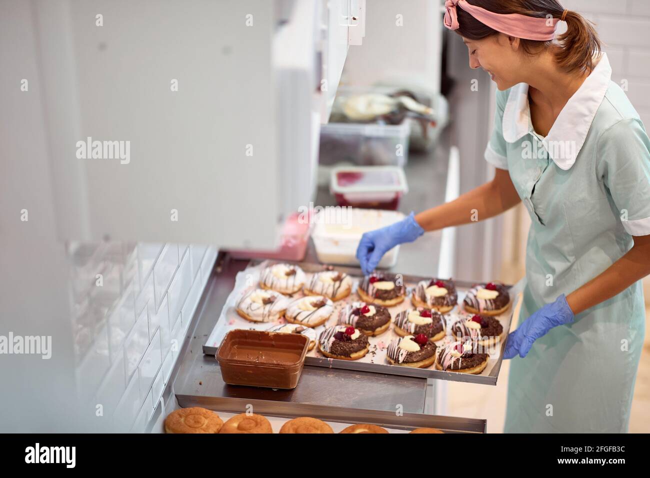 Una giovane piccola impresa donna proprietario sta decorando le ciambelle deliziose fatte a mano con una passione in un'atmosfera rilassata in un laboratorio. Pasticceria, dessert, swe Foto Stock