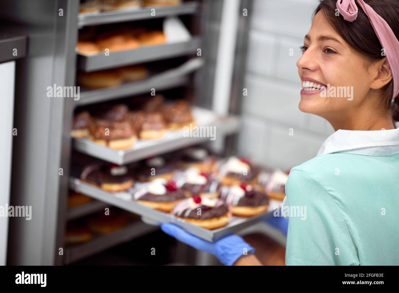 Una giovane piccola impresa donna proprietario in un'atmosfera piacevole in un laboratorio di caramelle che posa di fronte al guardaroba pieno di deliziose ciambelle fatte a mano. Passato Foto Stock