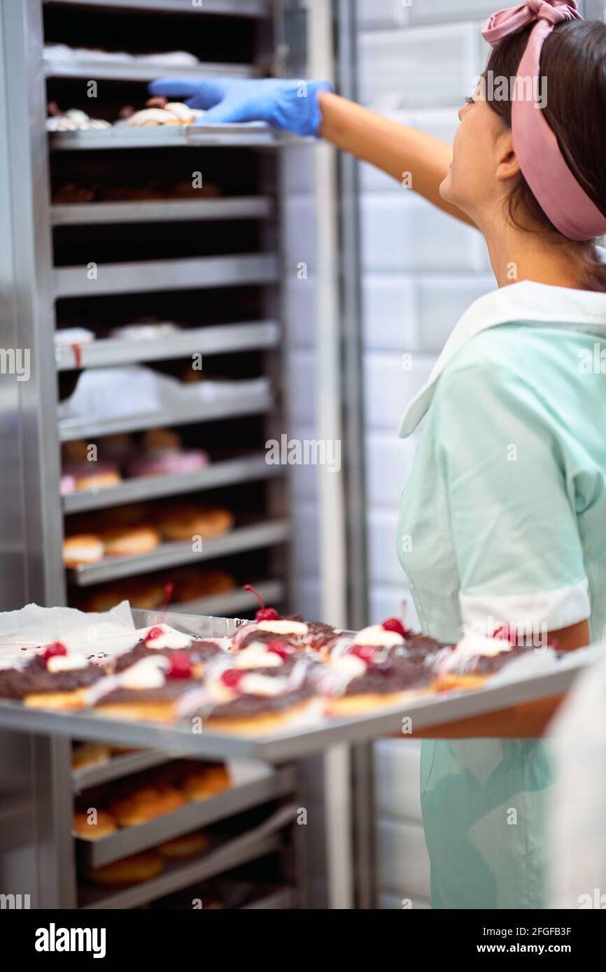Una giovane piccola impresa donna proprietario sta prendendo le ciambelle deliziose fatte a mano fuori da un owen in un'atmosfera di lavoro in un laboratorio caramelle. Pasticceria, dessert, Foto Stock