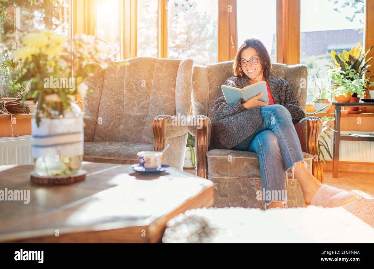 Allegra donna sorridente vestito accogliente abiti a casa seduta in comoda poltrona, rilassante, leggere un libro e bere caffè o tè del mattino in cous Foto Stock