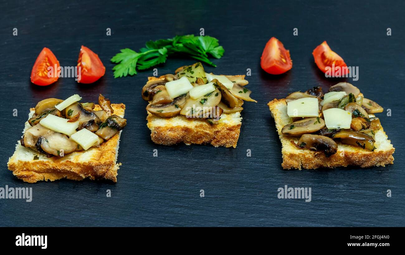 Tre fette di pane tostato con funghi champignon, prezzemolo, formaggio emmental e pomodori ciliegia rossi a fette, su piastra di ardesia Foto Stock