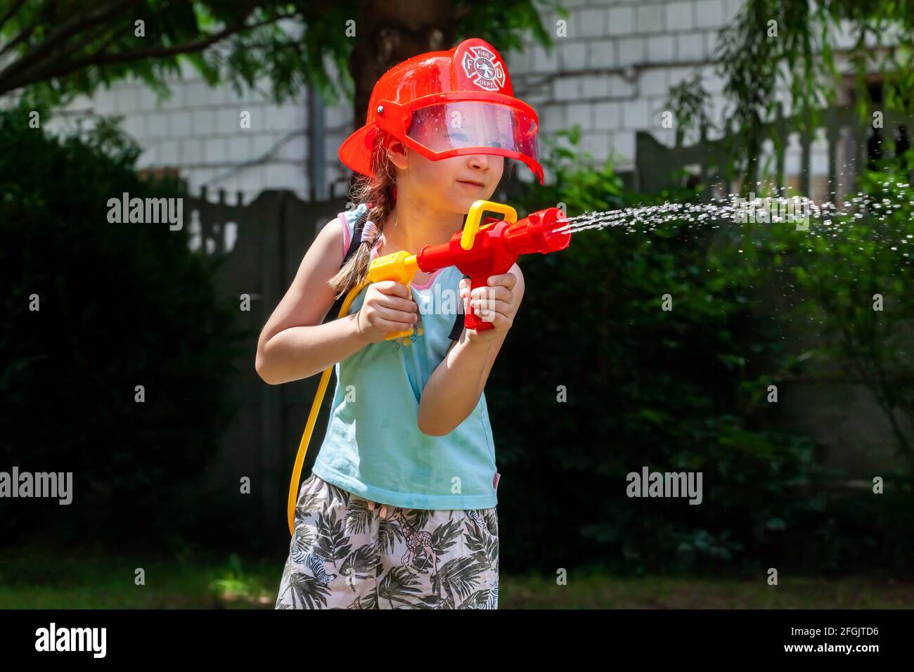 Pompiere, bambino, bambina in età scolare che indossa un casco da pompiere che gioca con una pistola ad acqua in giardino. Sogni, occupazione futura, lavoro, gioco Foto Stock
