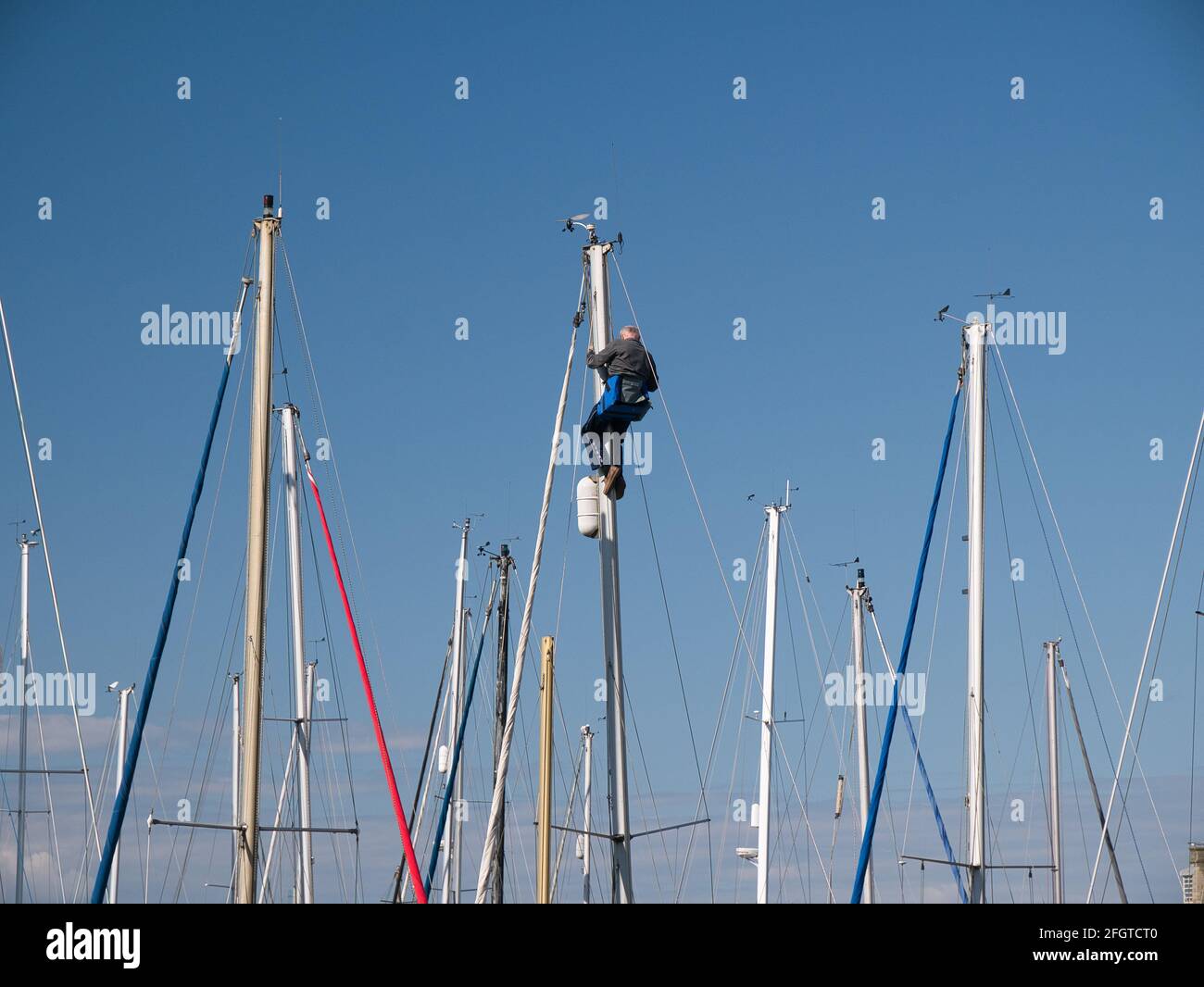 In una giornata tranquilla e soleggiata con un cielo blu chiaro, un uomo lavora in cima al palo di uno yacht ormeggiato in una marina. Appaiono altri montanti yacht. Foto Stock