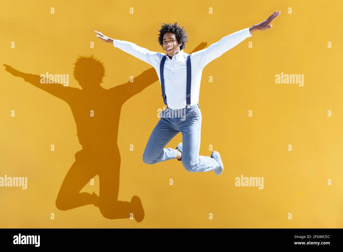 Uomo nero con capelli afro che saltano su uno sfondo urbano giallo. Ragazzo che indossa camicia e sospenditori. Foto Stock