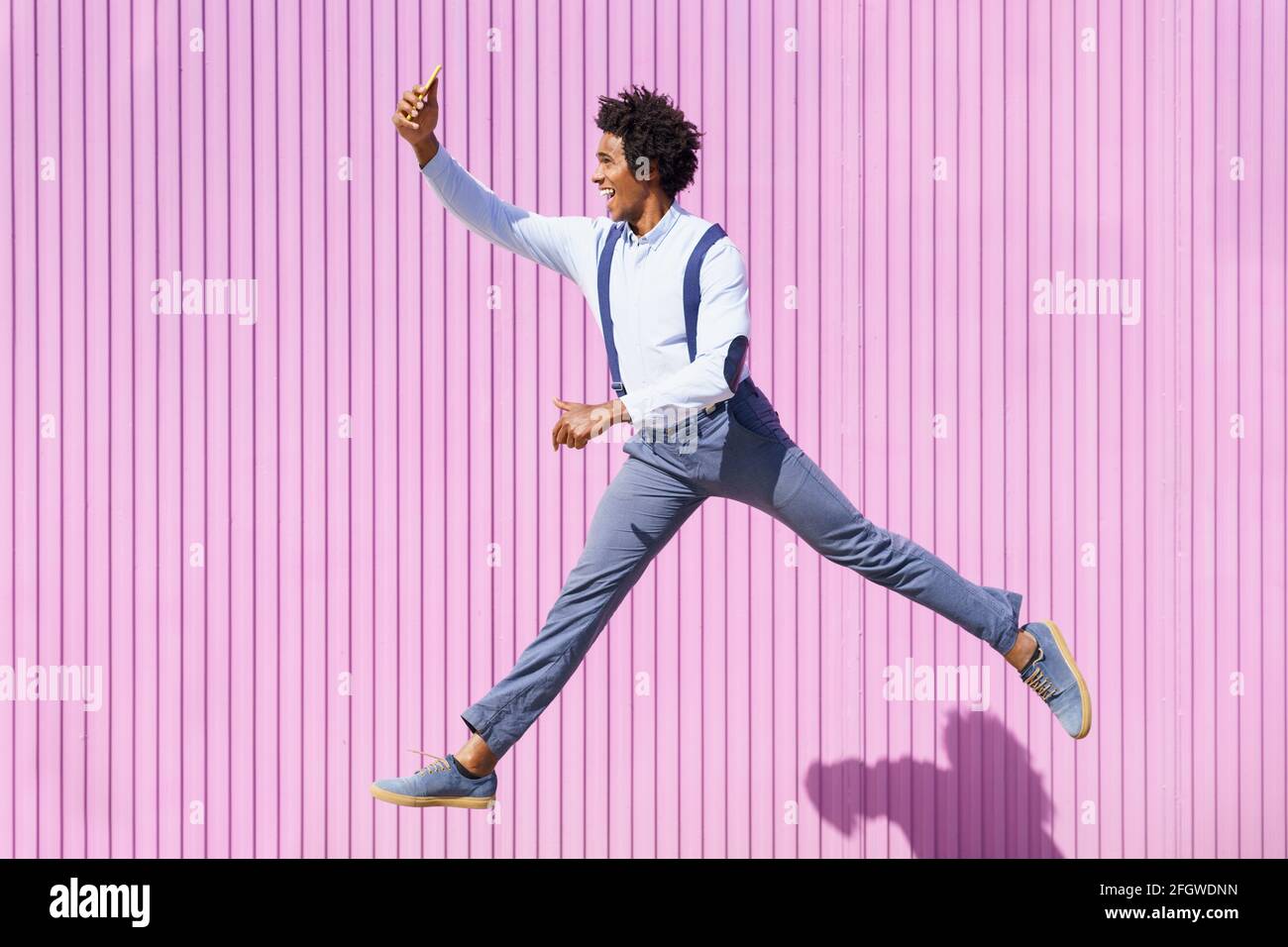 Uomo nero, con capelli afro, prendendo un selfie con il suo smartphone mentre saltando su uno sfondo di persiane rosa. Foto Stock