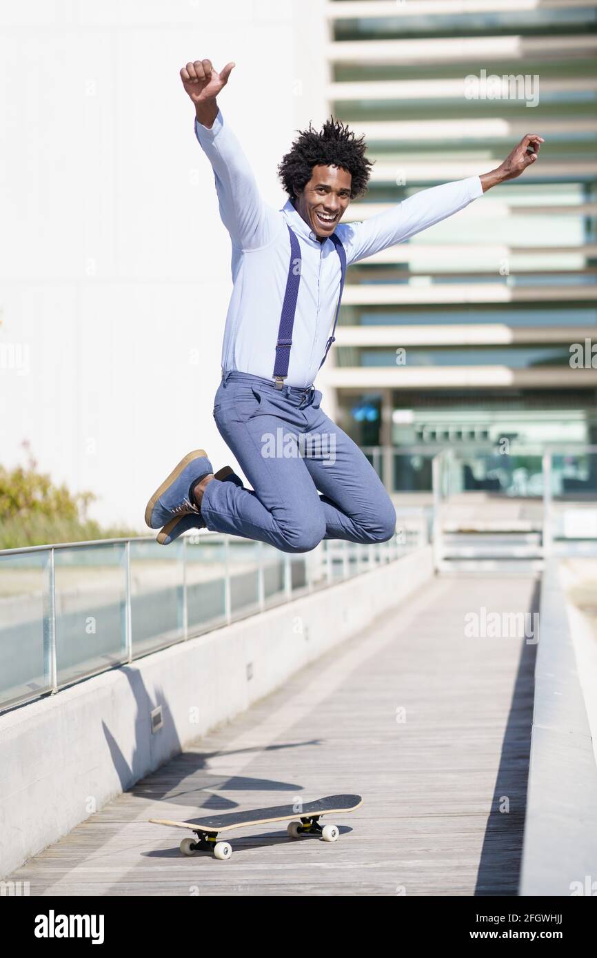 Uomo d'affari nero che salta su uno skateboard vicino a un edificio d'ufficio. Ragazzo con capelli afro. Foto Stock