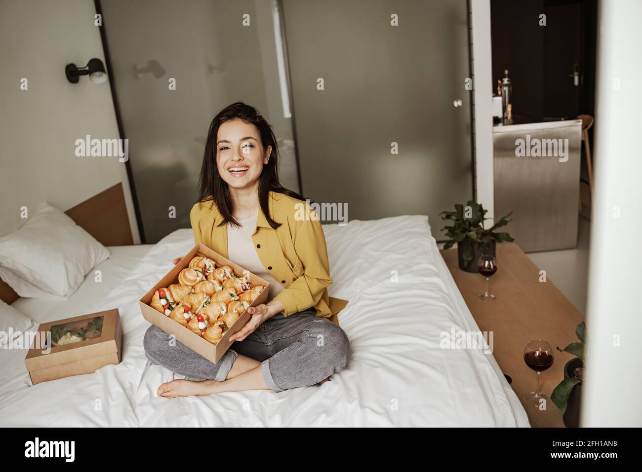 Bella donna seduta sul letto con scatole di cibo pieno di croissant, ha una cena romantica a casa Foto Stock