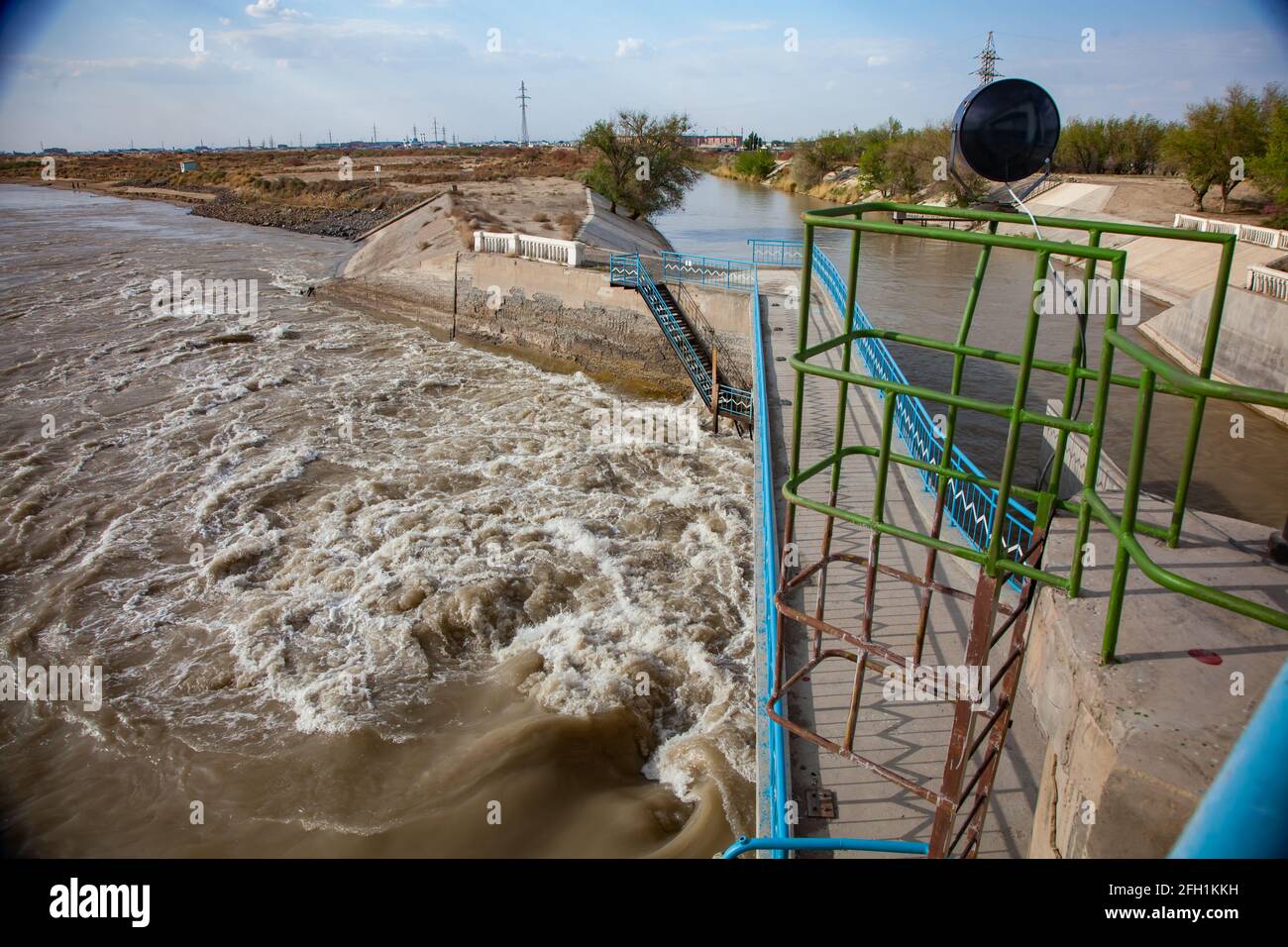 Diga del fiume Shardara con acqua gialla turbolenta, e canale a destra. Pareti di cemento. Balaustra in metallo, proiettore. Cielo blu. Kyzylorda, Kazakistan. Foto Stock