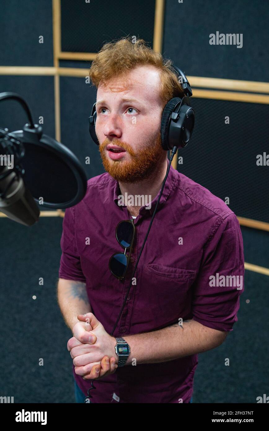 Vista laterale del giovane cantante maschile con cuffie che suonano la canzone vicino al microfono Foto Stock