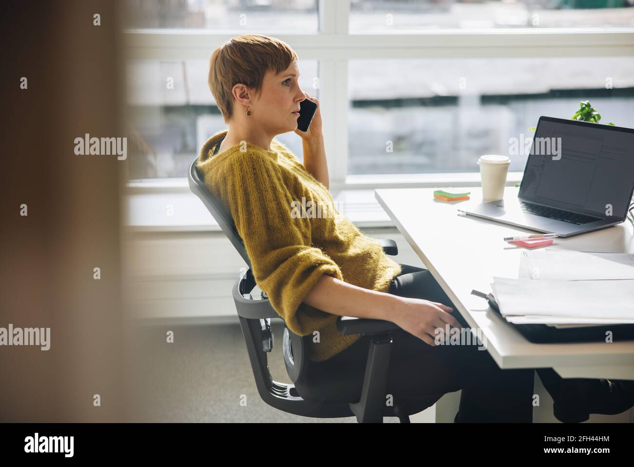 Vista laterale di una donna seduta alla scrivania che parla con il telefono cellulare. Lavoratore in ufficio che effettua una chiamata mentre lavora alla scrivania. Foto Stock