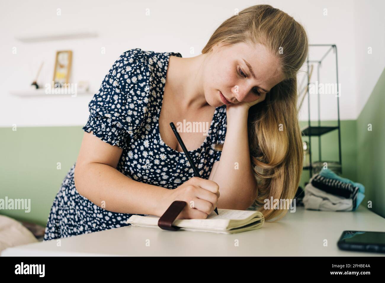 Giovane donna che si appoggia con la mano sulla scrivania mentre prende appunti in blocco note a casa alla luce del giorno Foto Stock