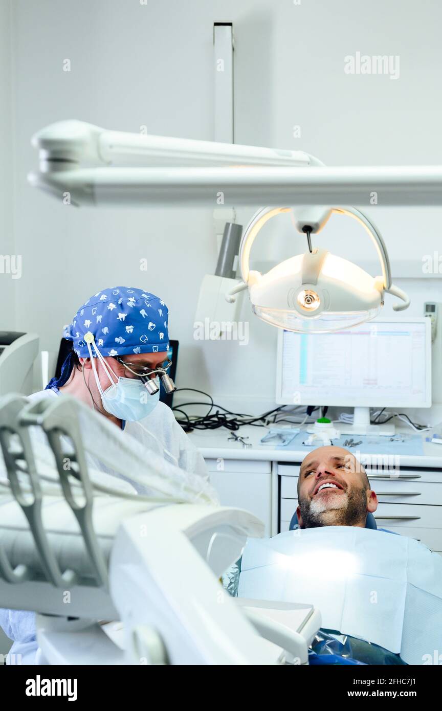 Vista laterale del chirurgo dentale anonimo in uniforme e medico binocoli con contenuto uomo sulla sedia in clinica Foto Stock