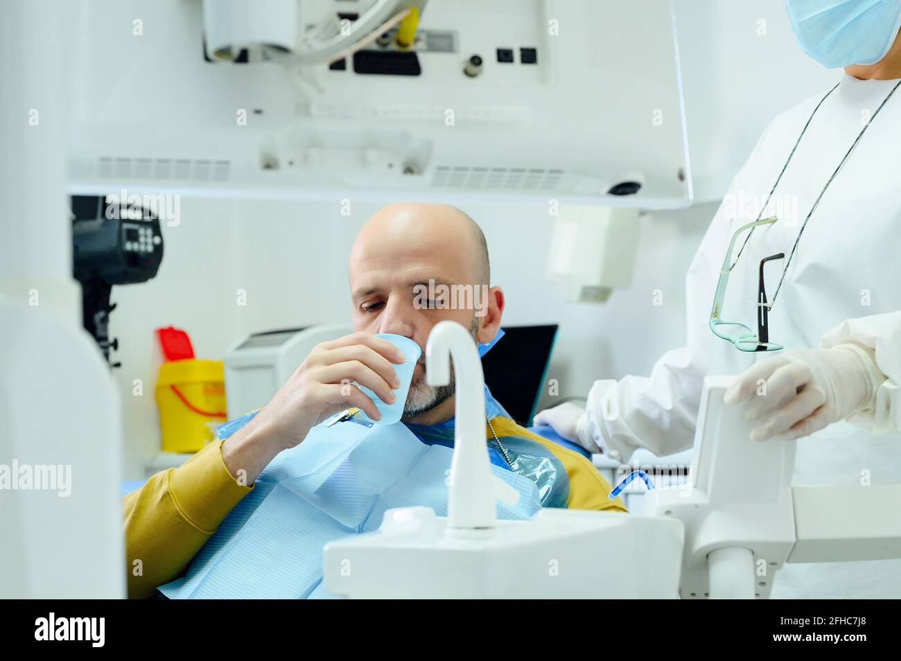 Coltivare dentista femminile non riconoscibile in maschera uniforme e sterile contro l'uomo versa l'acqua dal rubinetto in vetro dopo l'operazione orale Foto Stock