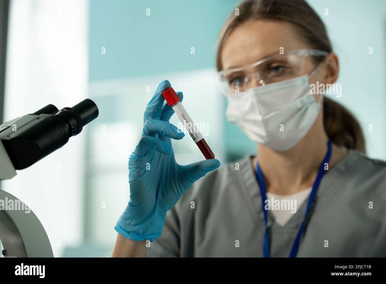 Primo piano di un giovane ricercatore di laboratorio in maschera e occhiali in piedi davanti al microscopio e tenendo un campione di sangue di coronavirus paziente Foto Stock