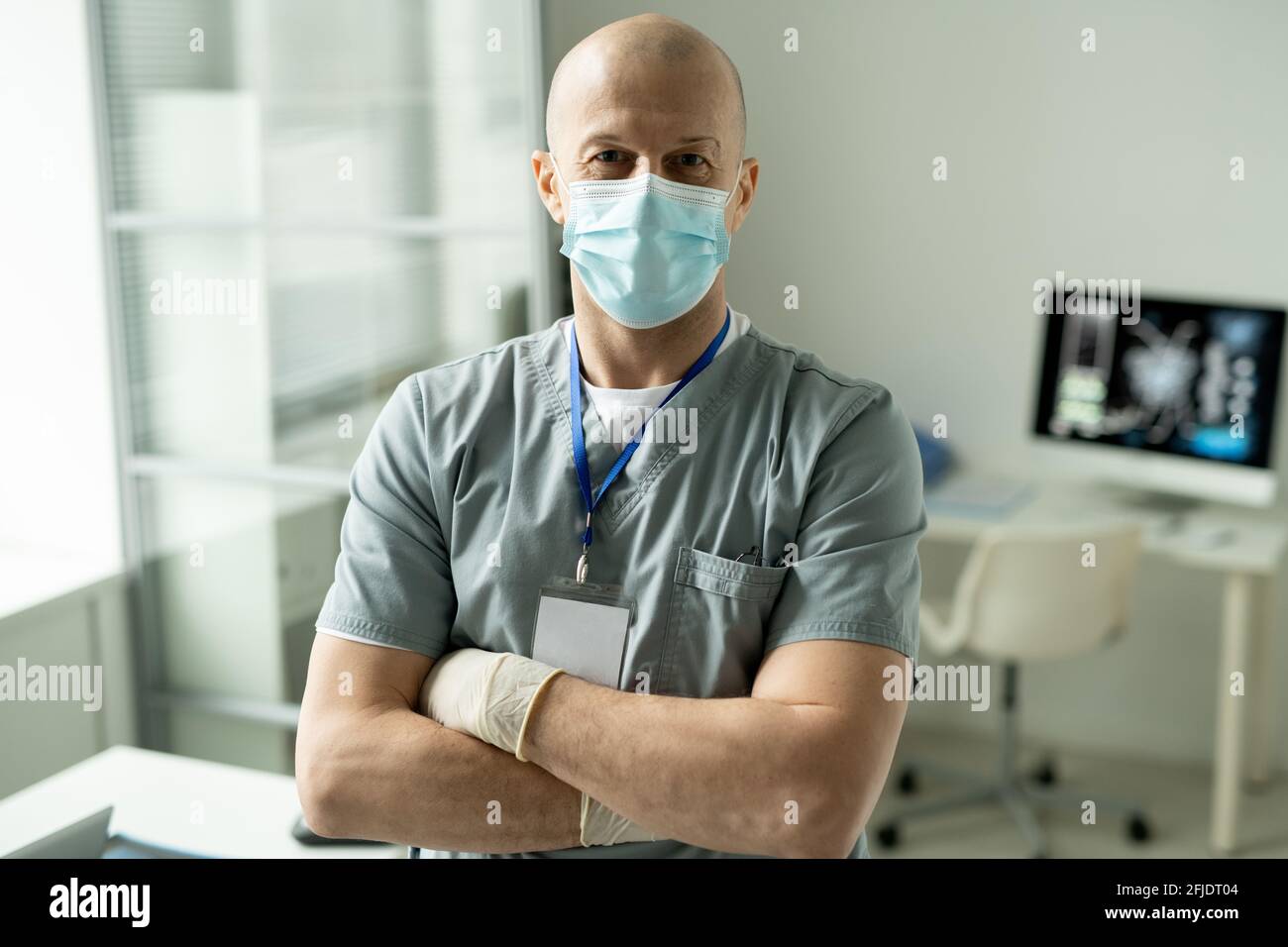 Ritratto del contenuto esperto medico specialista in maschera e scrub indossare il badge sul collo in piedi con le braccia incrociate in ufficio Foto Stock