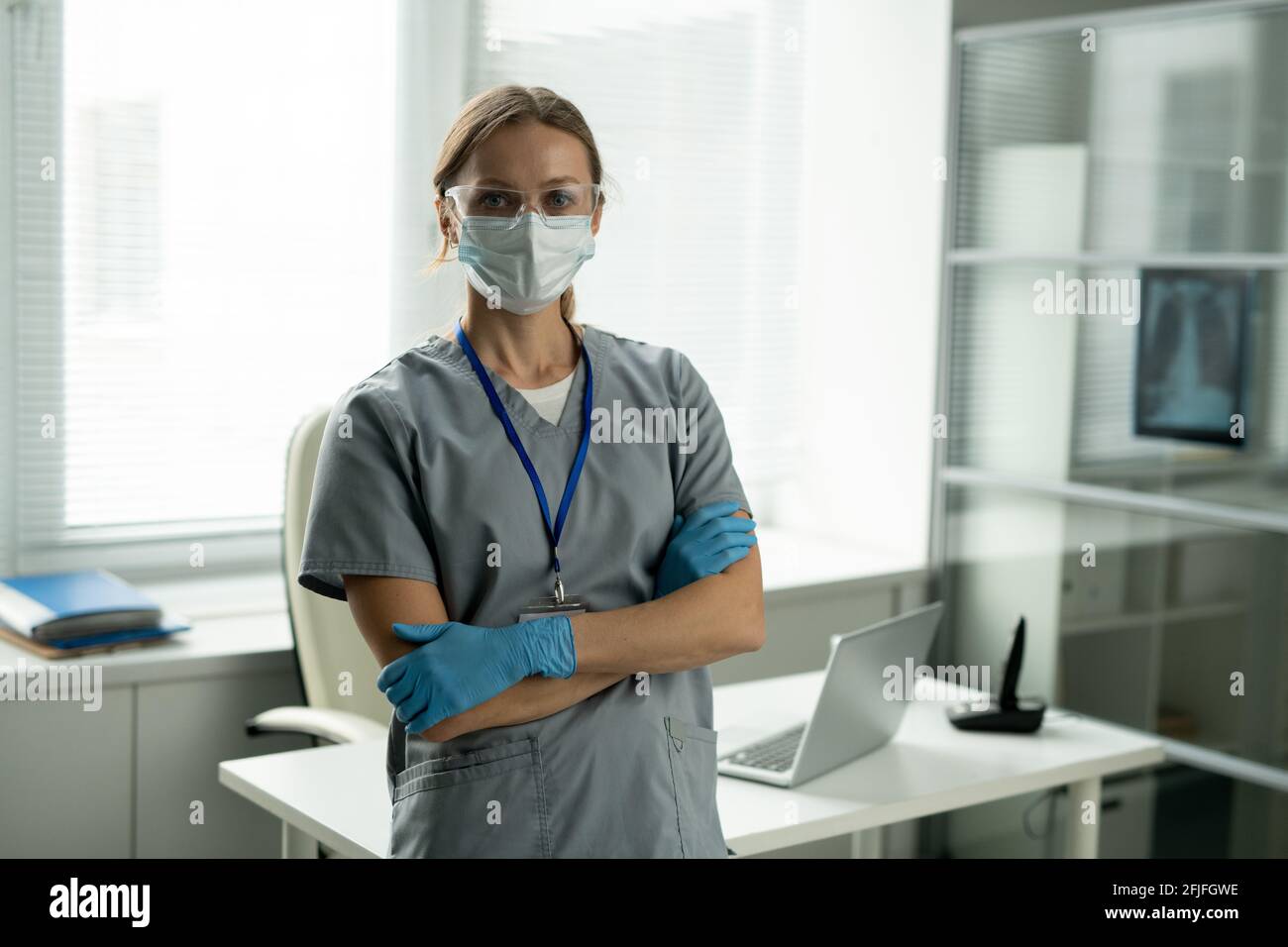 Ritratto di giovane chirurgo sicuro in maschera e guanti in piedi con braccia incrociate contro la scrivania nel proprio ufficio Foto Stock