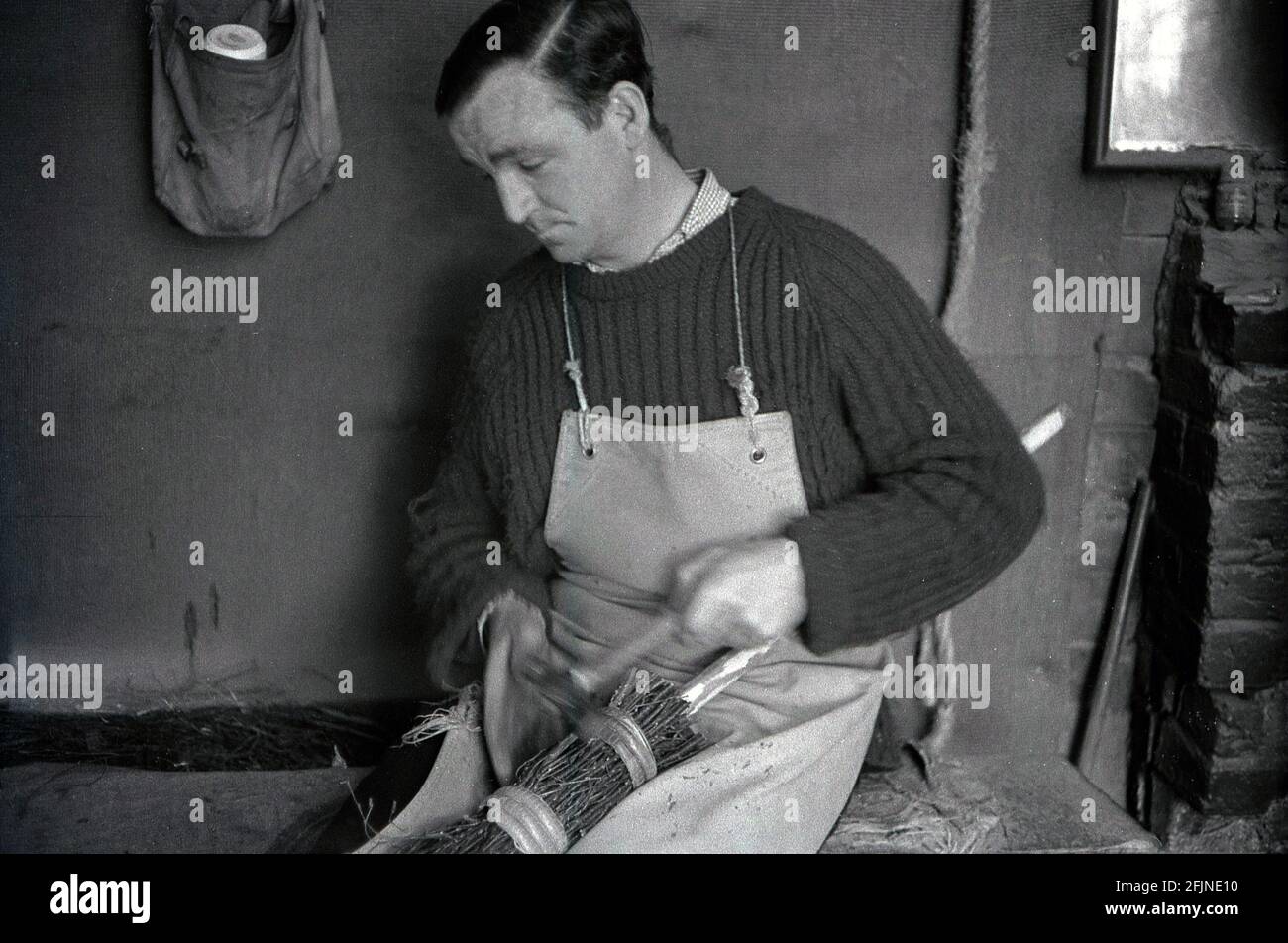 Anni '50, storico, un artigiano maschile nel suo laboratorio di fare una scopa, Inghilterra, Regno Unito. Un'antica imbarcazione, la realizzazione di scopa, conosciuta anche come 'besoms' o 'besom making', consiste in un fascio di ramoscelli di alberi legati saldamente intorno ad un bastone. Tradizionalmente, le scope besom erano fatte da ramoscelli dell'albero della betulla e uno squire della scopa del villaggio ed entrambi i coppiceworker li hanno prodotti. Oggi, come molti antichi mestieri, questo mestiere altamente specializzato è quasi completamente morto fuori e ci sono ora meno di cinque creatori di besom nel Regno Unito che fanno le scopa su una base commmerica. Foto Stock