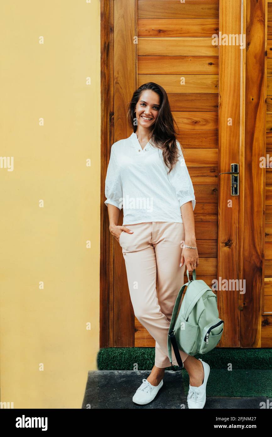 Giovane ragazza elegante in una blusa bianca, pantaloni color crema, sneaker bianche e con uno zaino verde sullo sfondo di una parete gialla e di un woo Foto Stock