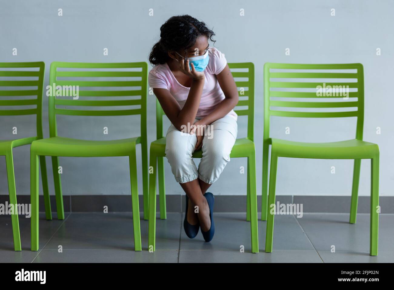 Misto razza ragazza che indossa maschera di faccia seduta su sedia verde in sala d'attesa ospedaliera Foto Stock