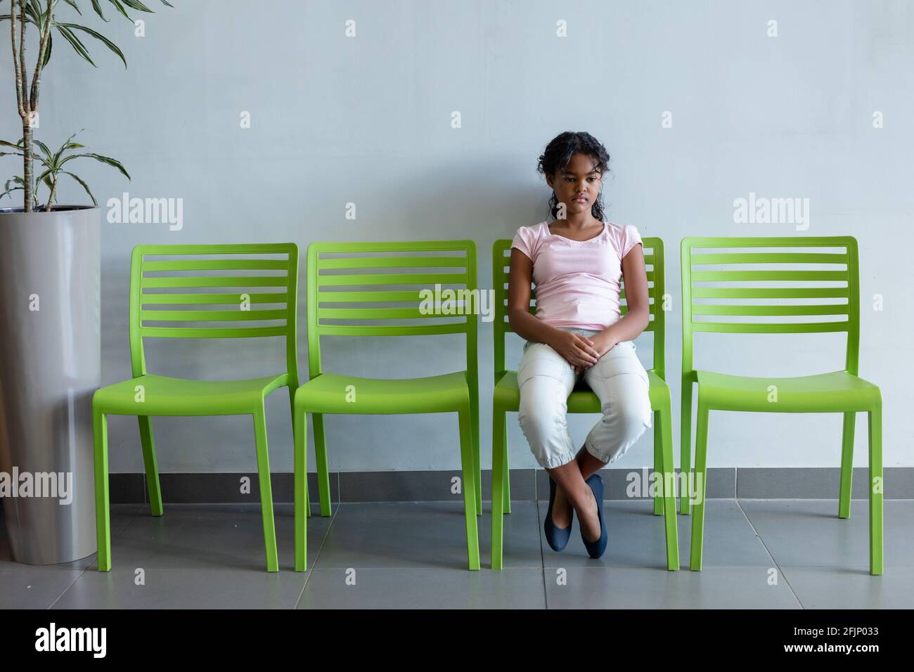Annoiata ragazza mista seduta su sedia verde in ospedale sala d'attesa Foto Stock