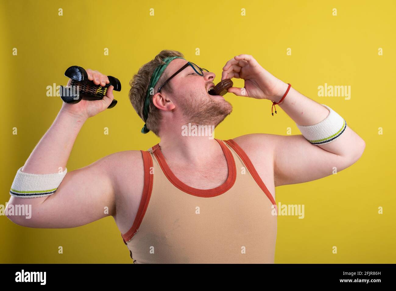 divertente uomo grasso in abiti sportivi con manubri nel suo mani mangia biscotti al cioccolato Foto Stock