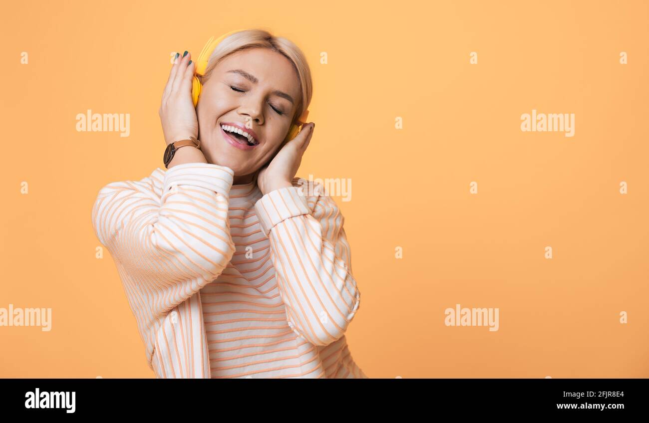 Una donna affascinante sta ascoltando la musica sulle cuffie che si posano una parete gialla dello studio che fa pubblicità a qualcosa Foto Stock