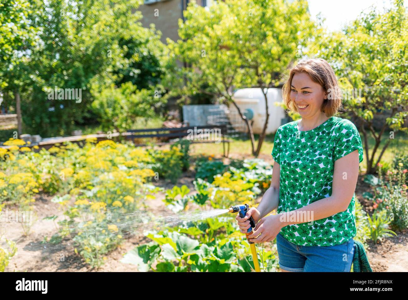 Una giovane donna caucasica allegra infondendo i letti con il verde con un tubo.Estate. Il concetto di giardinaggio e ricreazione estiva. Foto Stock