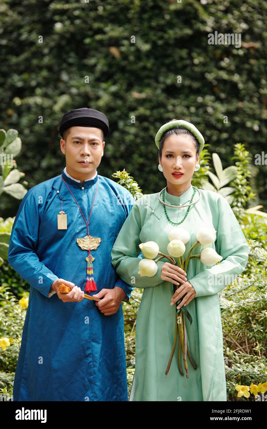 Ritratto di una giovane coppia vietnamita preoccupata in costumi tradizionali situato nel parco cittadino con fiori di loto e fan della carta Foto Stock
