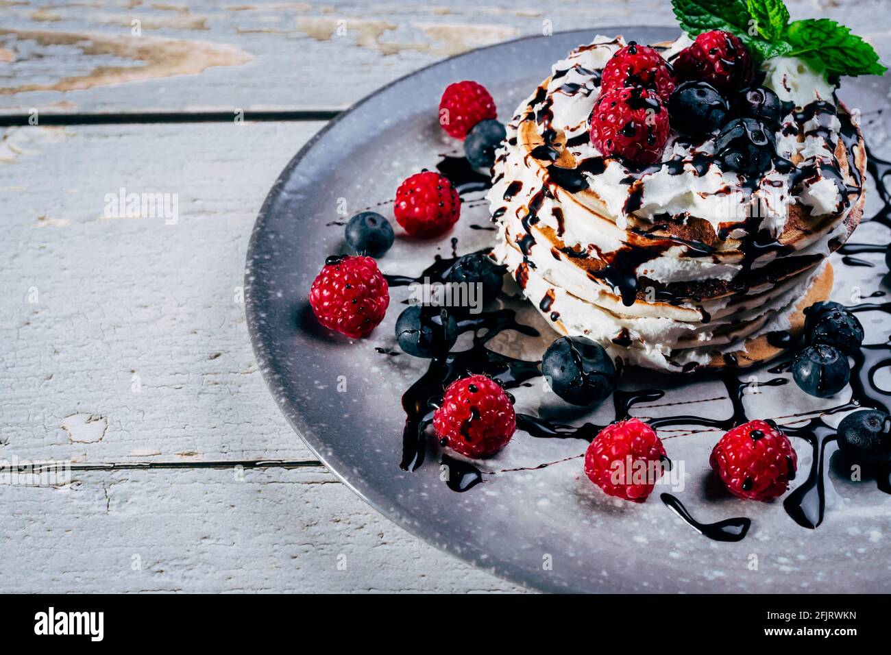piatto di frittelle con crema e sciroppo di cioccolato con mirtilli, fragole e lamponi su rustico tavolo di legno. Foto Stock