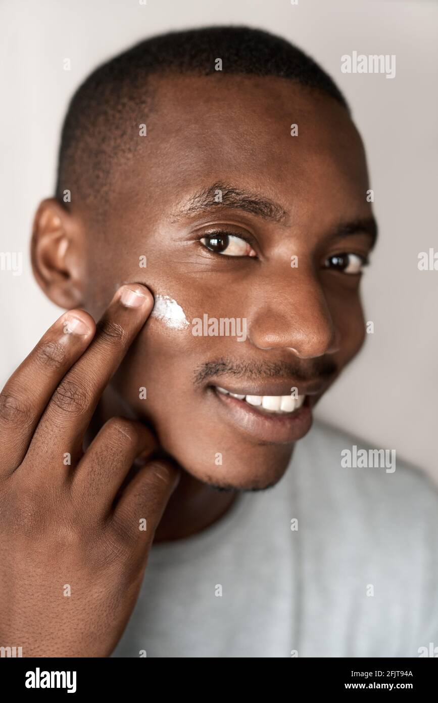 Sorridente giovane africano che applica crema idratante alla guancia Foto Stock