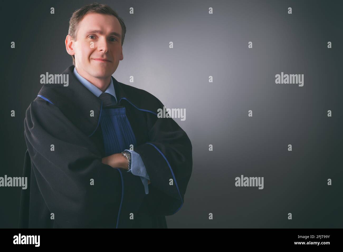 Un avvocato sorridente in una toga si alza con le braccia piegate su uno sfondo scuro Foto Stock