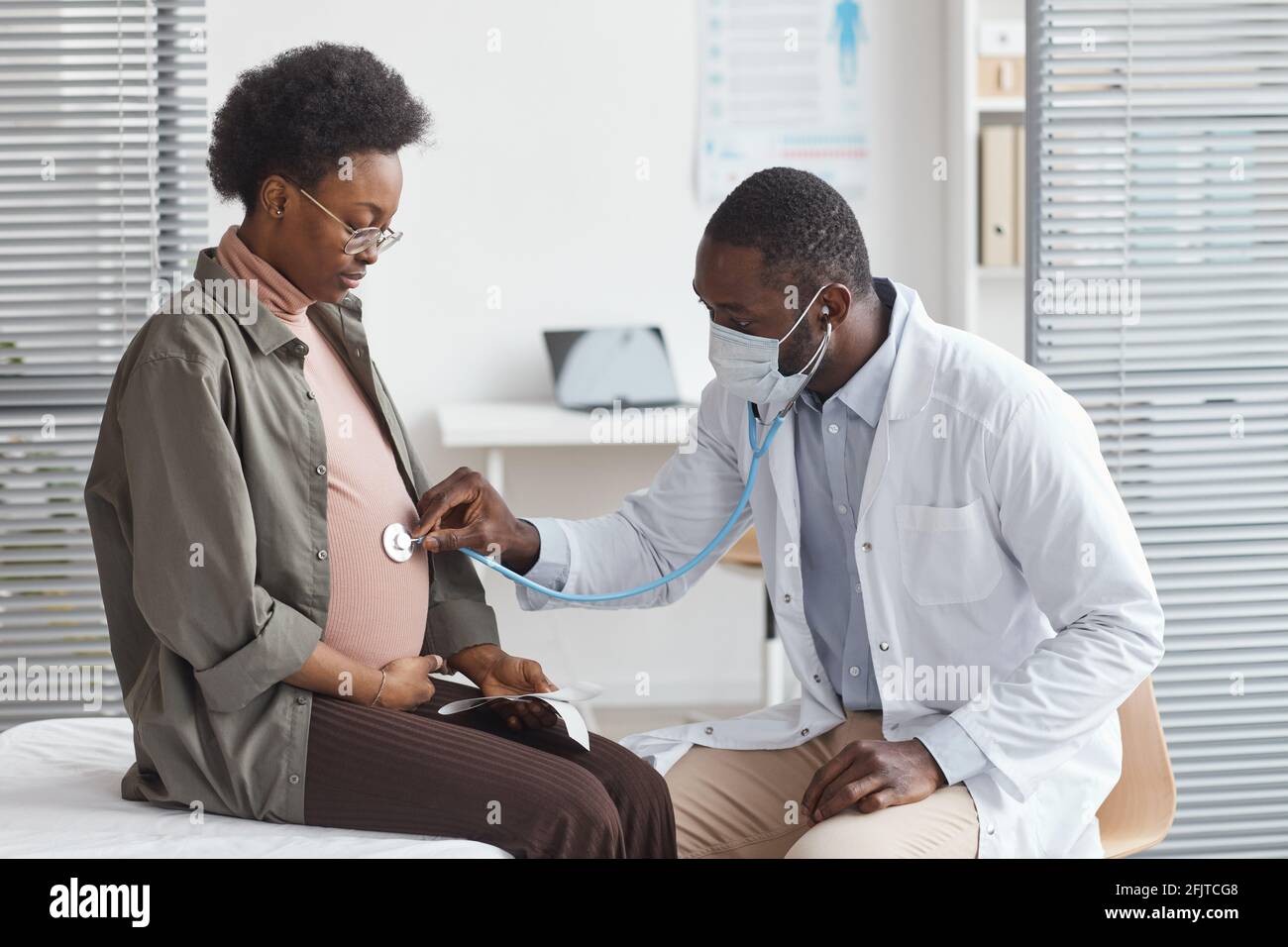 Medico africano in camice bianco che esamina la pancia di incinta donna con stetoscopio durante la visita in ospedale Foto Stock