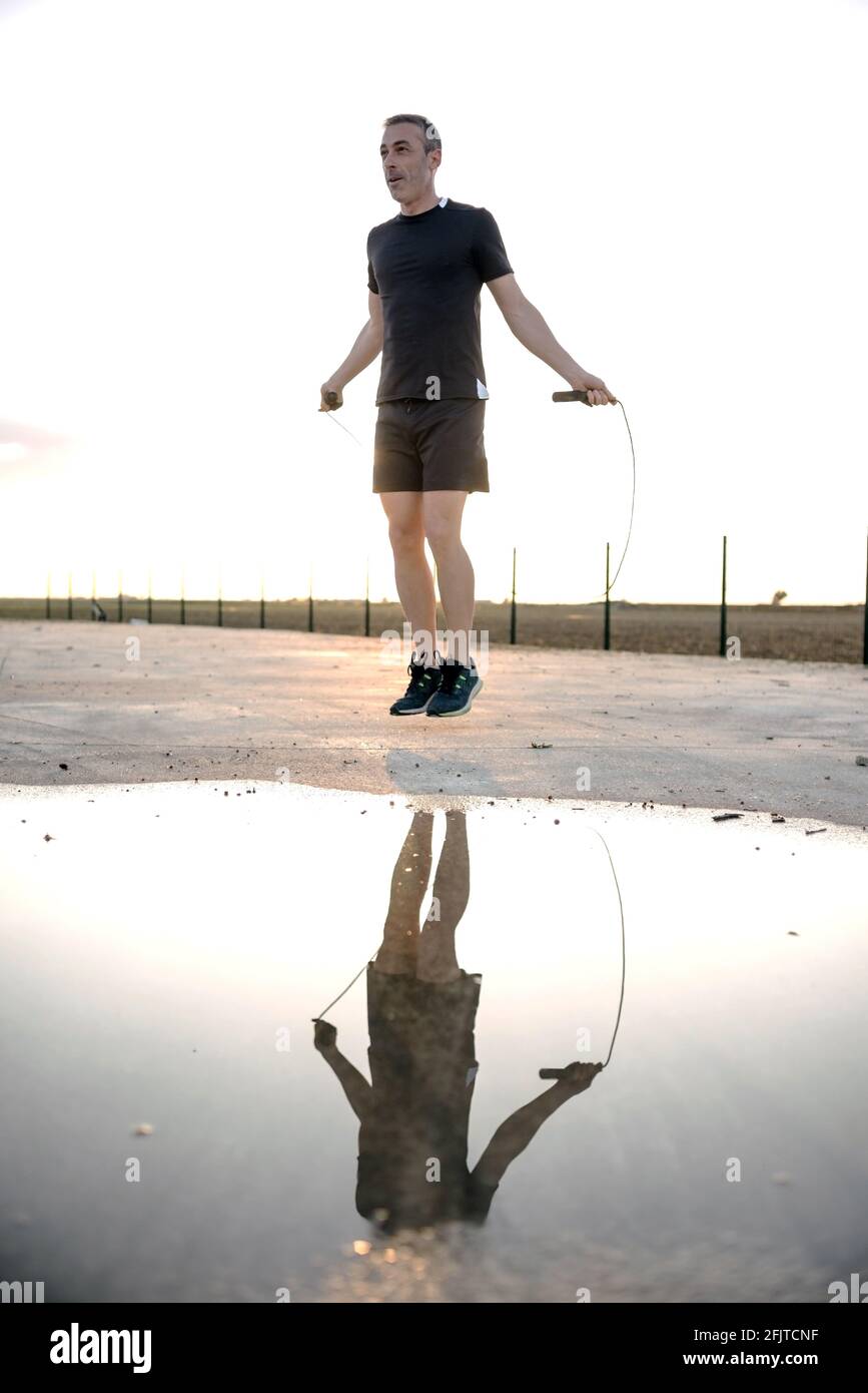 Concetto: sport. Atleta uomo all'aperto. Corda che salta. Retroilluminazione al tramonto. Riflesso nell'acqua di una pozza. Foto Stock