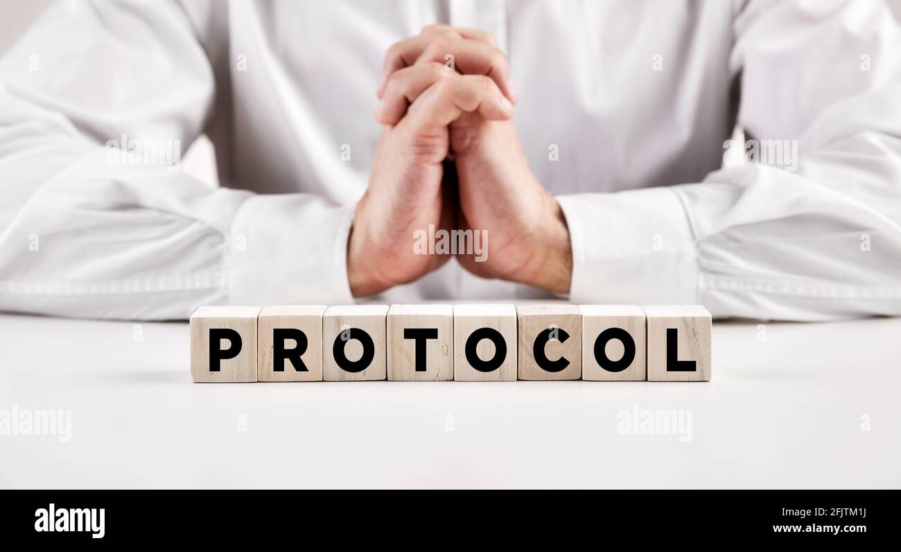 La parola protocollo scritto su un blocco di legno di fronte ad un uomo d'affari con le mani incrociate. Protocollo aziendale, procedure, regole o concetto di standard. Foto Stock