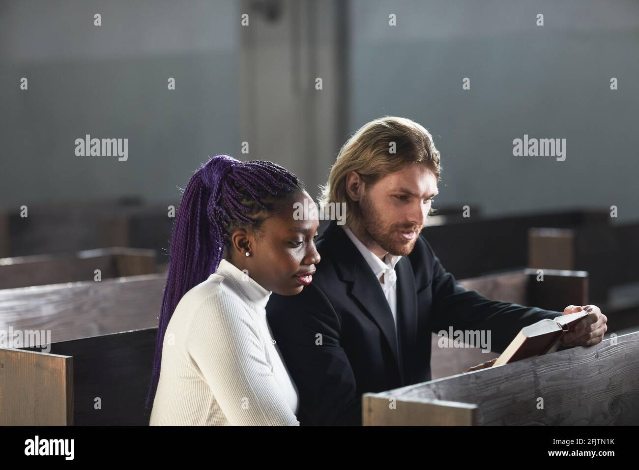 Giovane coppia multietnica seduta sulla panchina insieme e leggendo La Bibbia insieme nella chiesa Foto Stock
