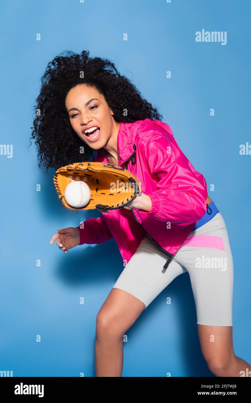 entusiasta donna afroamericana in guanto di pelle che cattura il baseball sopra blu Foto Stock