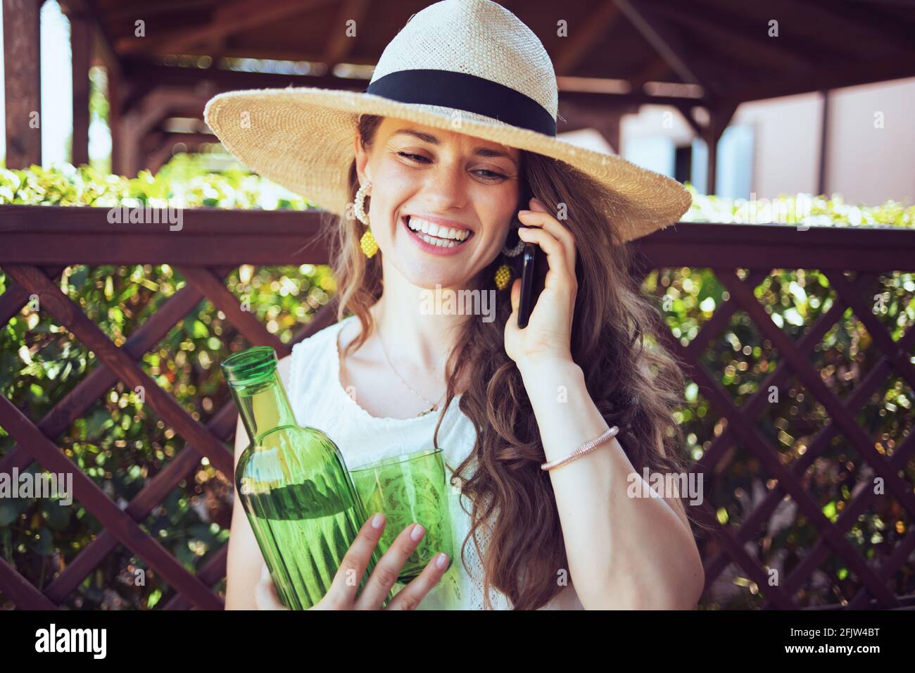 sorridente elegante donna di mezza età in camicia bianca con bottiglia d'acqua, vetro e cappello utilizzando uno smartphone in terrazza. Foto Stock