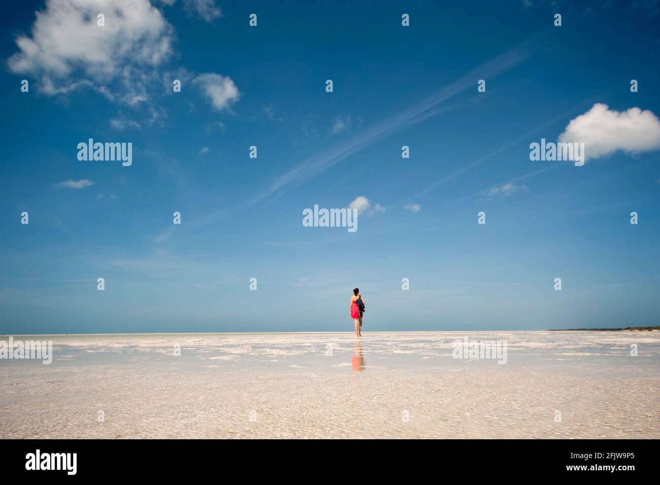 Retrovisione di una donna in un vestito rosso che cammina sulla spiaggia sull'isola di Holbox in Messico a bassa marea. Sullo sfondo l'orizzonte sul mare e bluer Foto Stock